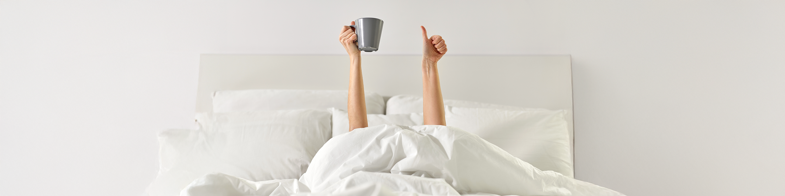 Arms holding a coffee mug above cozy white bedding, representing a relaxed and intentional morning vibe