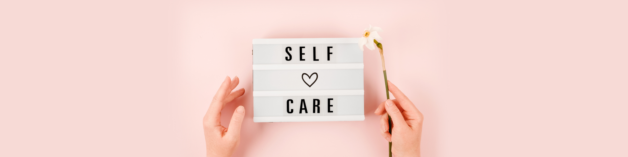 Hands holding a ‘Self Care’ lightbox sign on a soft pink background, representing mindful routines.
