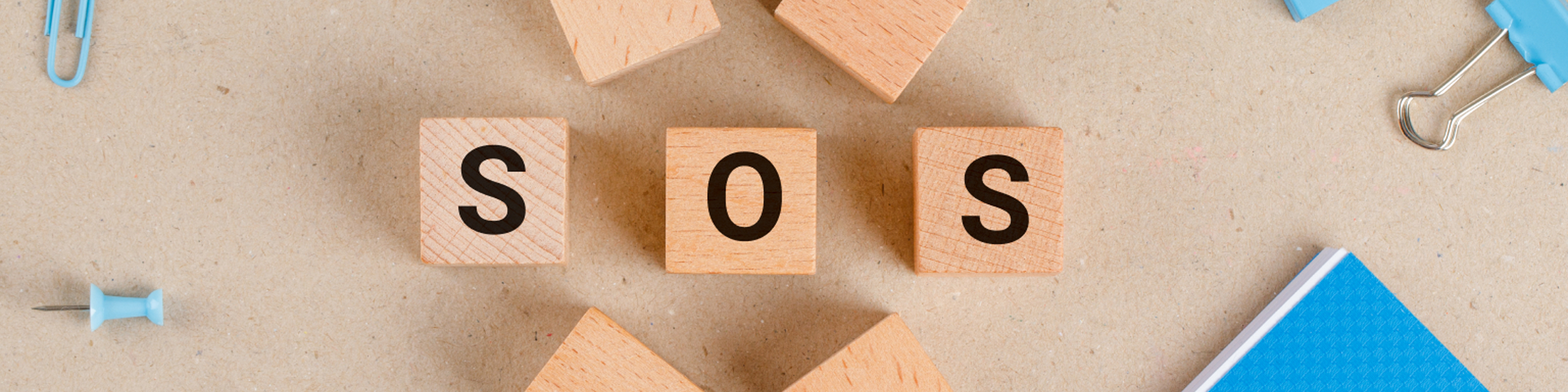 Wooden blocks spelling âSOSâ surrounded by office supplies, conveying stress and the need for routine rescue.