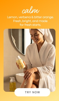 Woman in robe and towel on head sitting on bathroom counter applying Dear Me Calm body lotion next to Calm body wash smiling in a bright relaxing post-shower moment